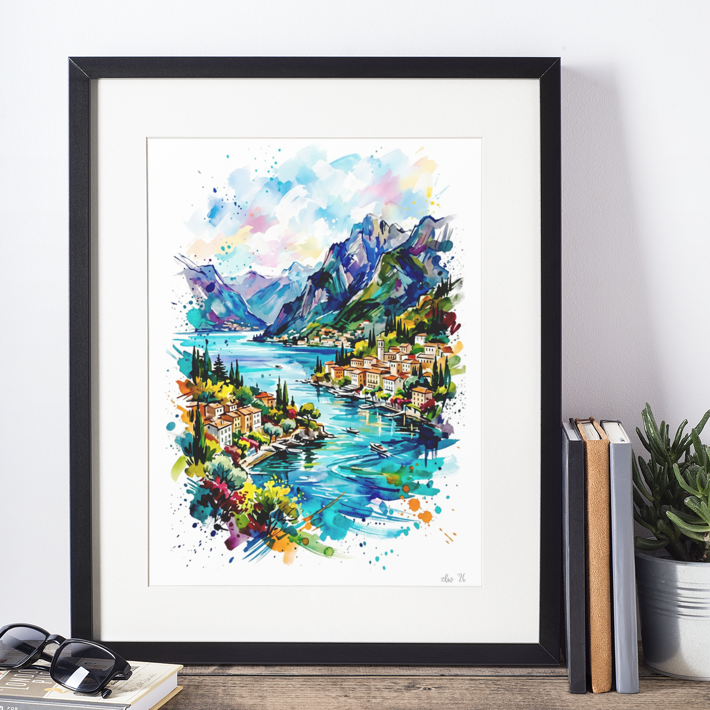 A framed watercolor art print depicting a colorful mountain lake town scene, styled on a wooden shelf with books, sunglasses, and a potted plant against a white wall.