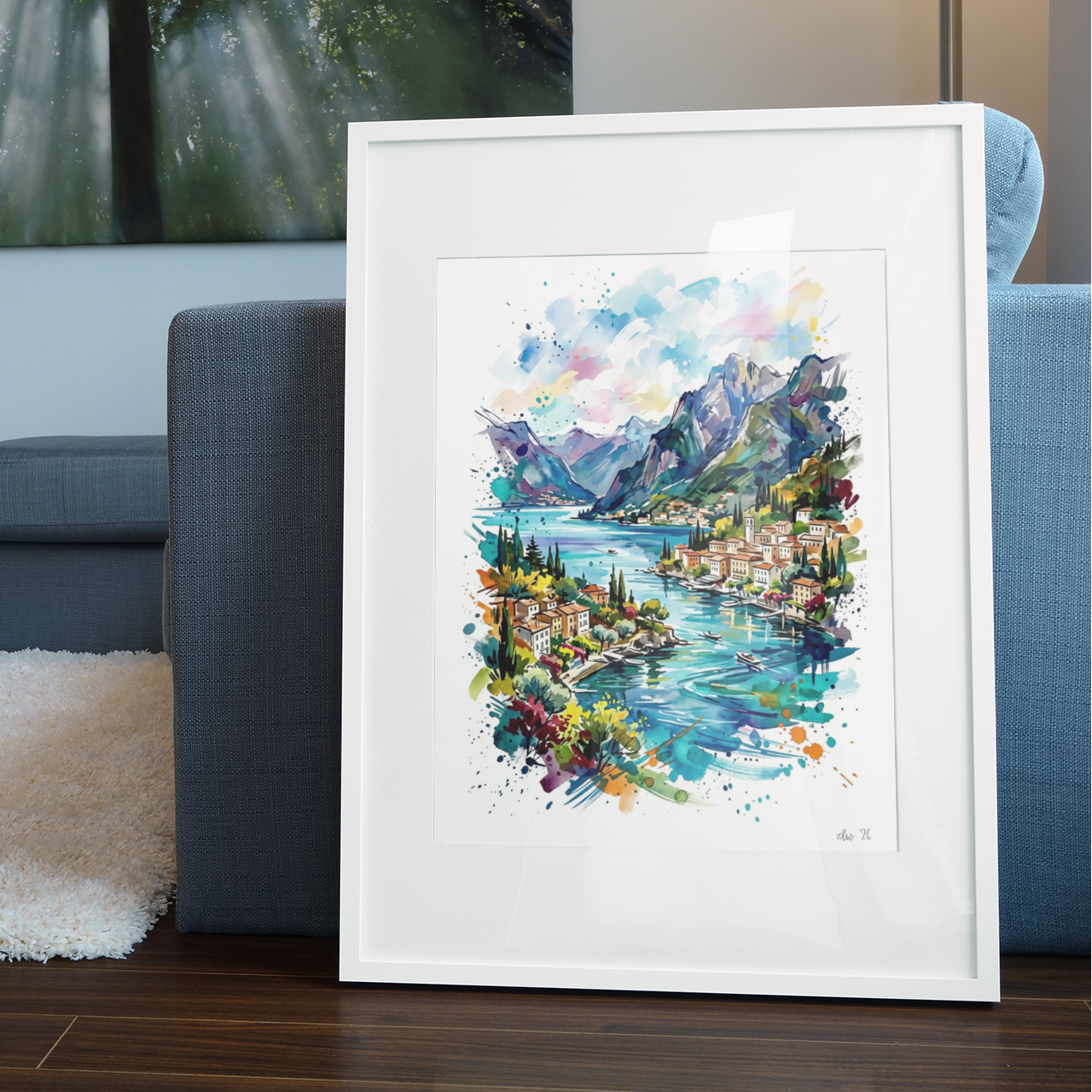 A framed watercolor art print depicting a scenic mountain lake landscape with colorful buildings and trees, leaning against a blue sofa in a modern living room setting.